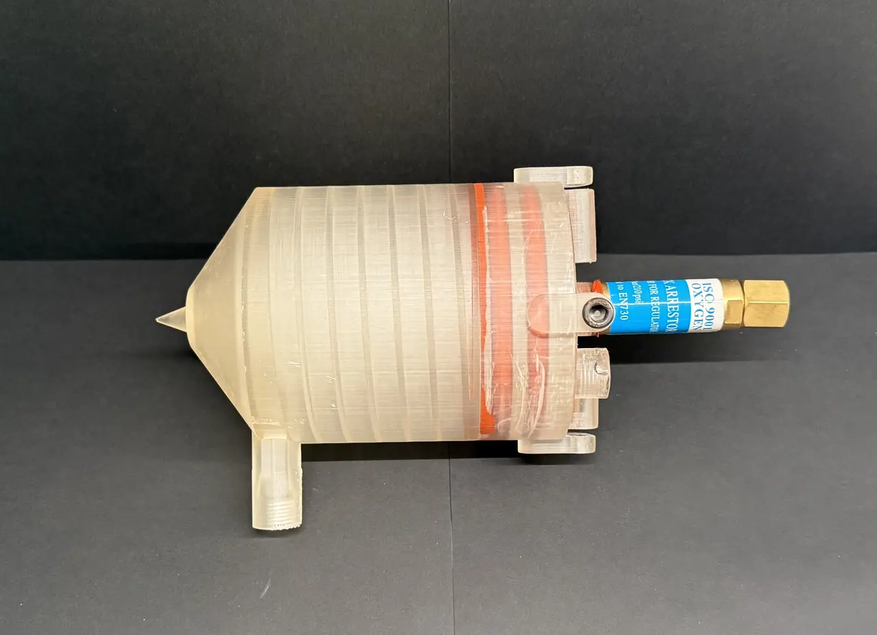 Translucent resin-printed aerospike engine lying on a dark surface with a conical spike nose, an orange seal ring and a blue-labelled brass fitting